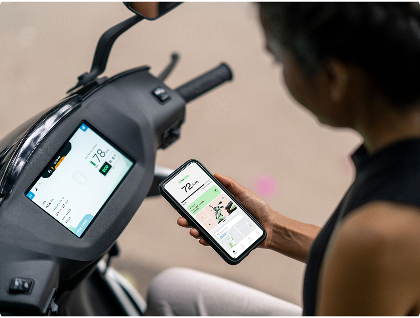 A rider on her scooter holds up a phone showing the Ather app, beside the scooter’s dashboard