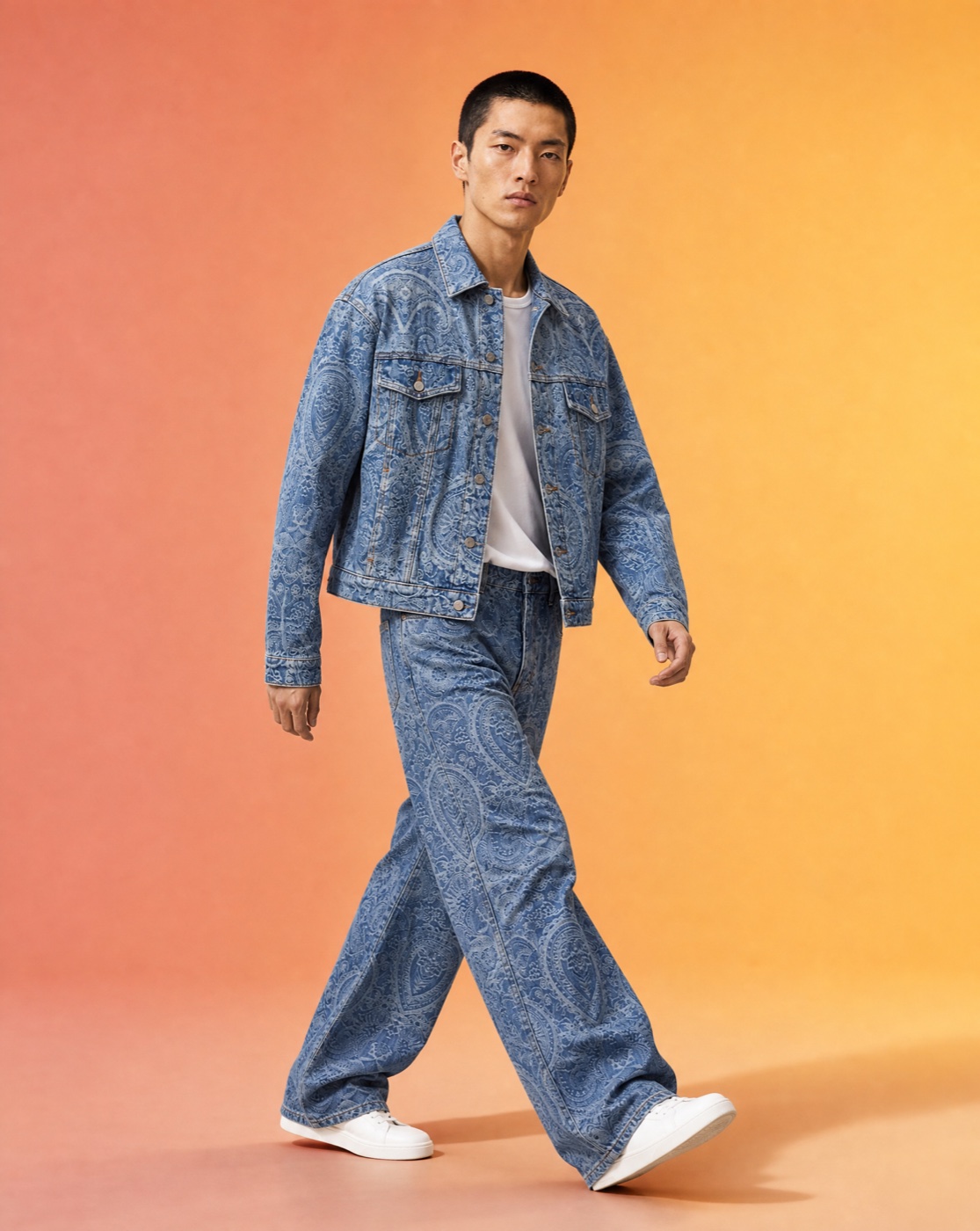 Model walking mid-stride in a blue paisley denim jacket and matching trousers in a warm studio.