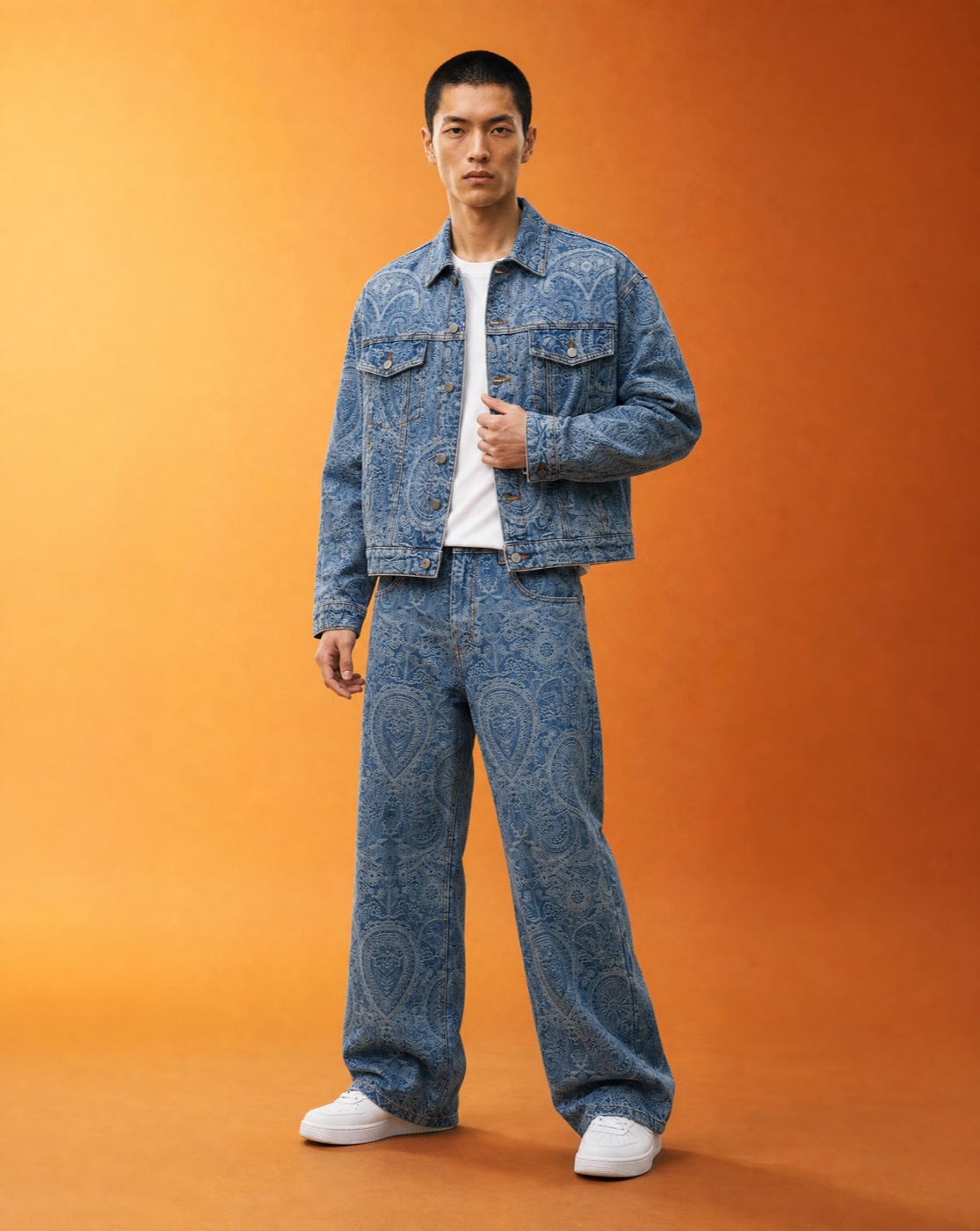 Model standing straight on holding a blue paisley denim jacket, with matching trousers, in a warm studio.