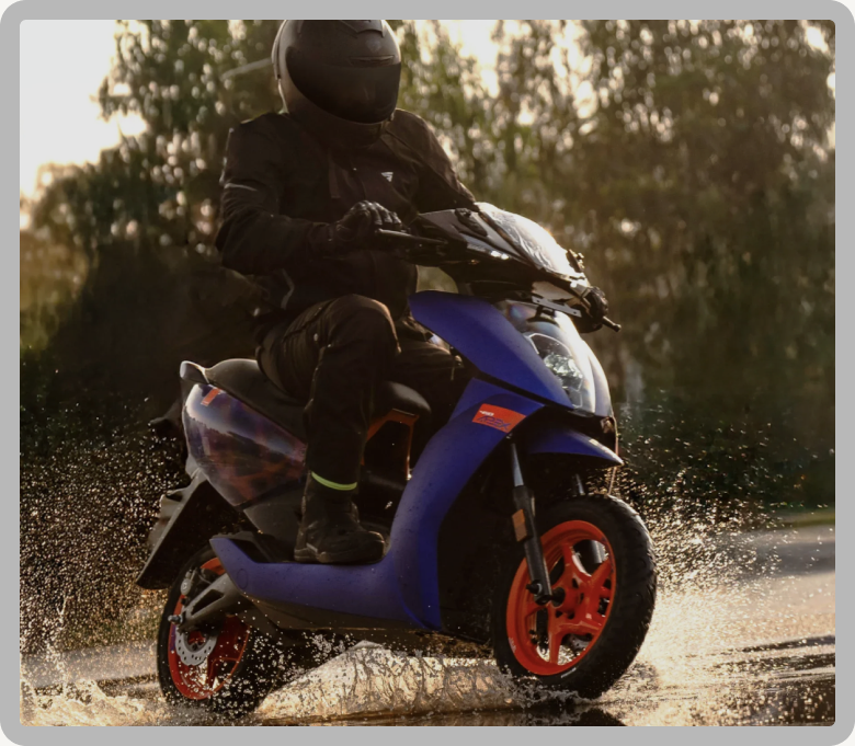 A rider in black gear on a blue Ather scooter splashing through water, with trees in warm evening light behind