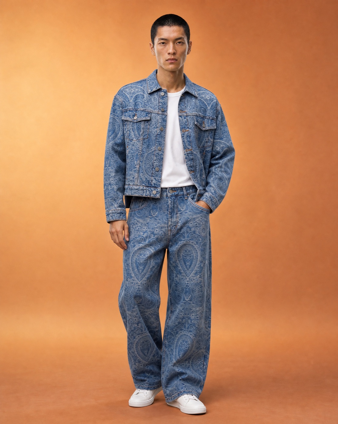 Model standing straight on with one hand in the pocket in a blue paisley denim jacket and matching trousers in a warm studio.