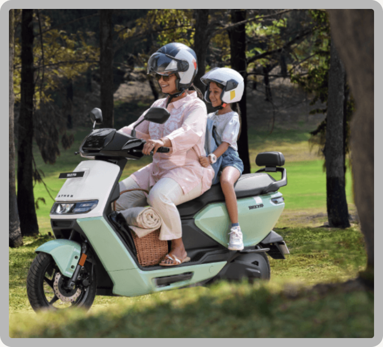 A woman and a child in helmets riding a pale green Ather scooter through a park