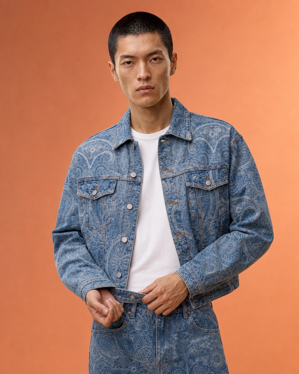 Chest-up studio portrait of the model adjusting a blue paisley denim jacket over a white t-shirt in a warm studio.