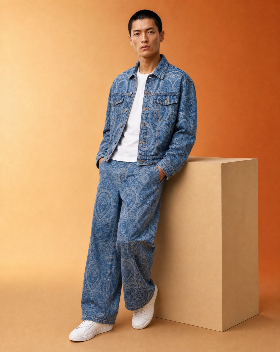 Model leaning against a pale block in a blue paisley denim jacket and matching trousers in a warm studio.
