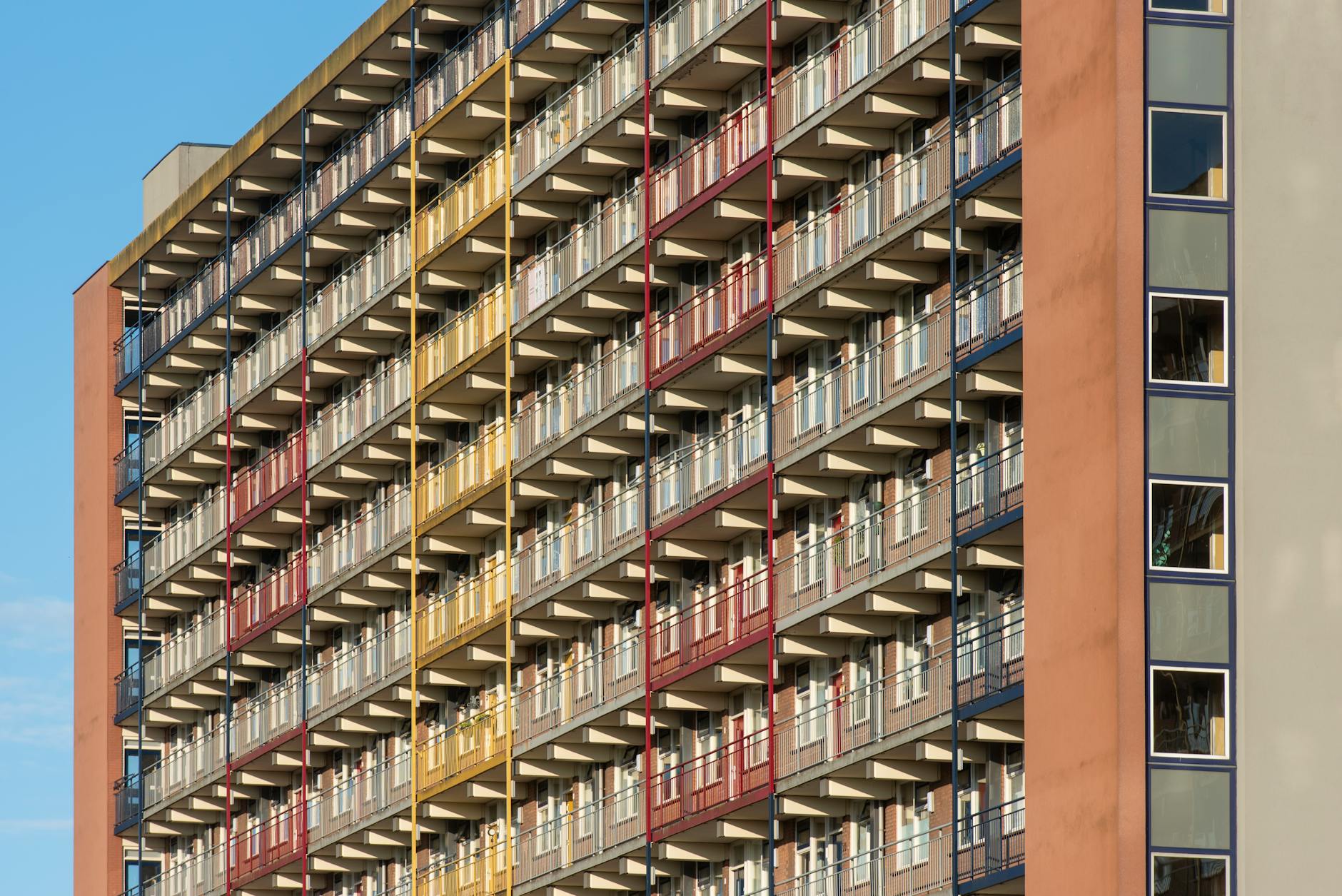 Tall residential block with rows of balconies and red and yellow painted railings on a terracotta facade against a blue sky