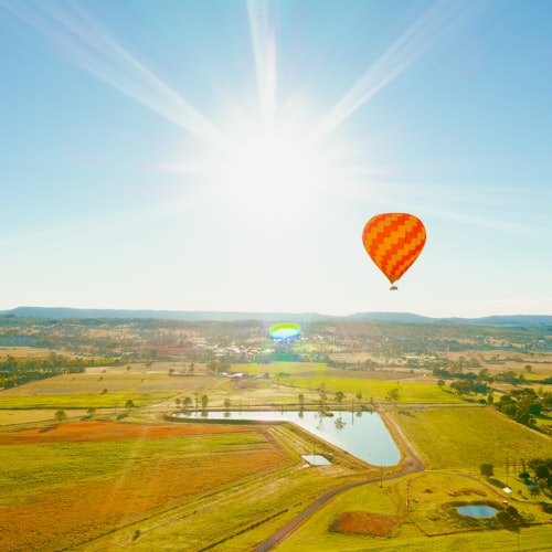 Drie heteluchtballonnen, één grote gele met de tekst "Austra", en twee kleinere, zweven boven een schilderachtig landschap met heuvels en bomen.