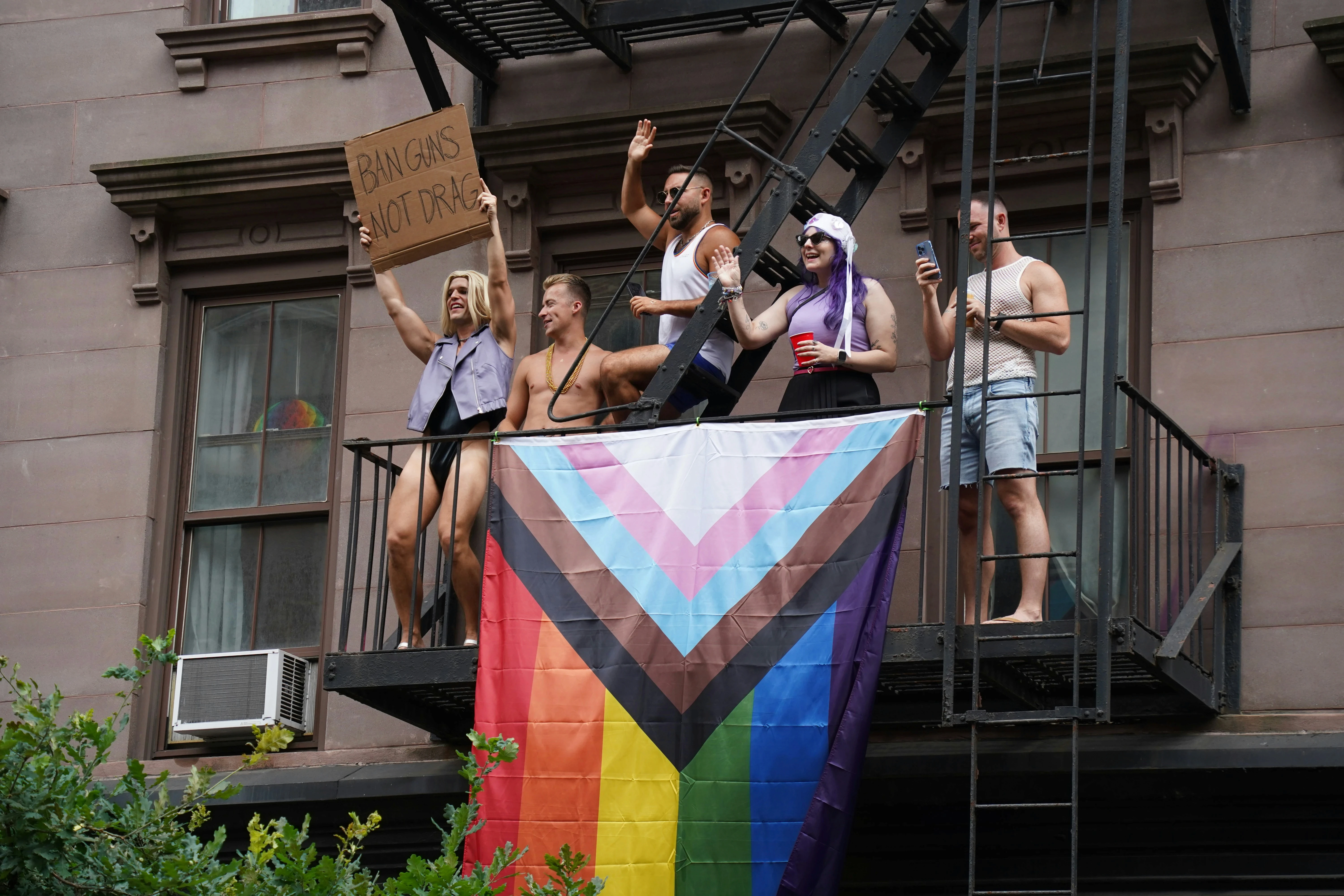 LGBTQ+ community members displaying a Progress Pride flag and protest sign from a fire escape during a public demonstration, symbolizing queer activism, solidarity, and collective resilience.