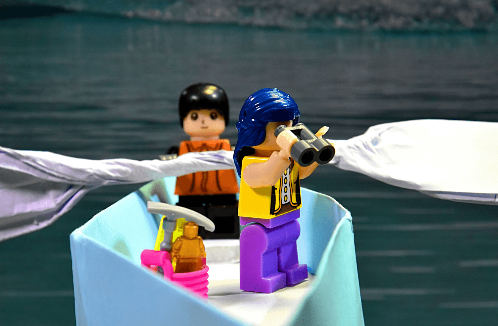 two lego characters in a paper boat, on a body of water. One character is looking to the left, while the other is looking to the right with a set of binoculars