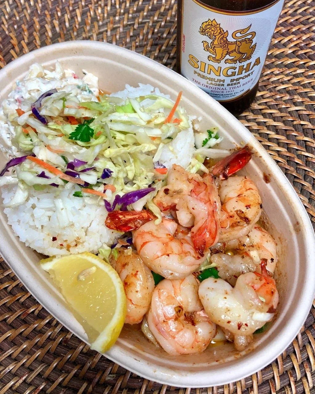 A bowl of buttery garlic shrimp garnished with red chili flakes and green herbs, served with two scoops of rice, macaroni salad, Asian slaw, and a lemon wedge.