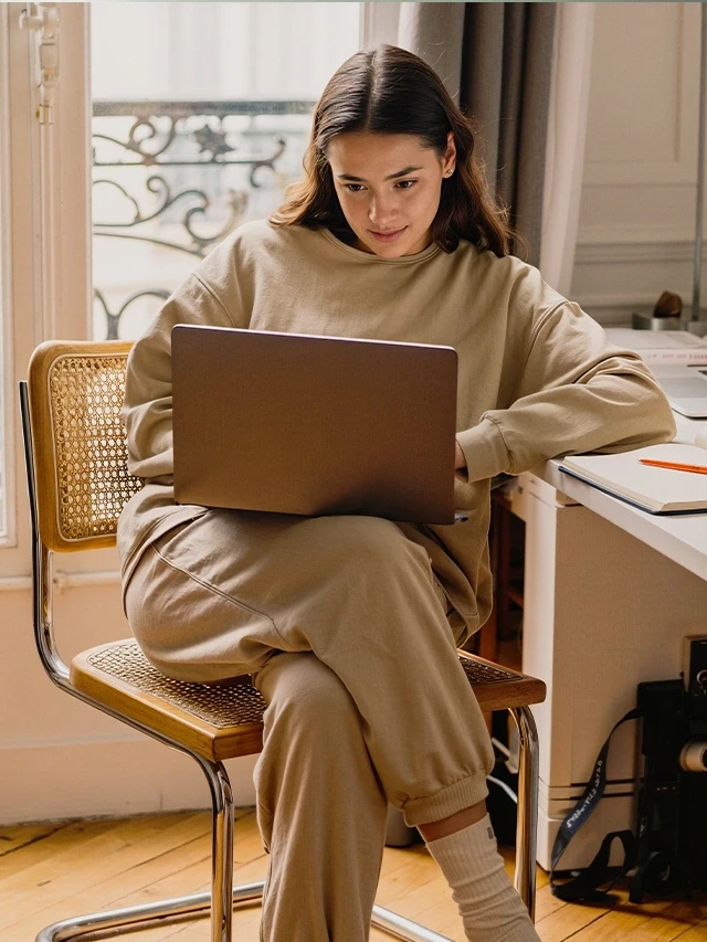 Woman in a beige outfit working on a laptop at an organized desk in a bright, cozy, naturally lit room. 