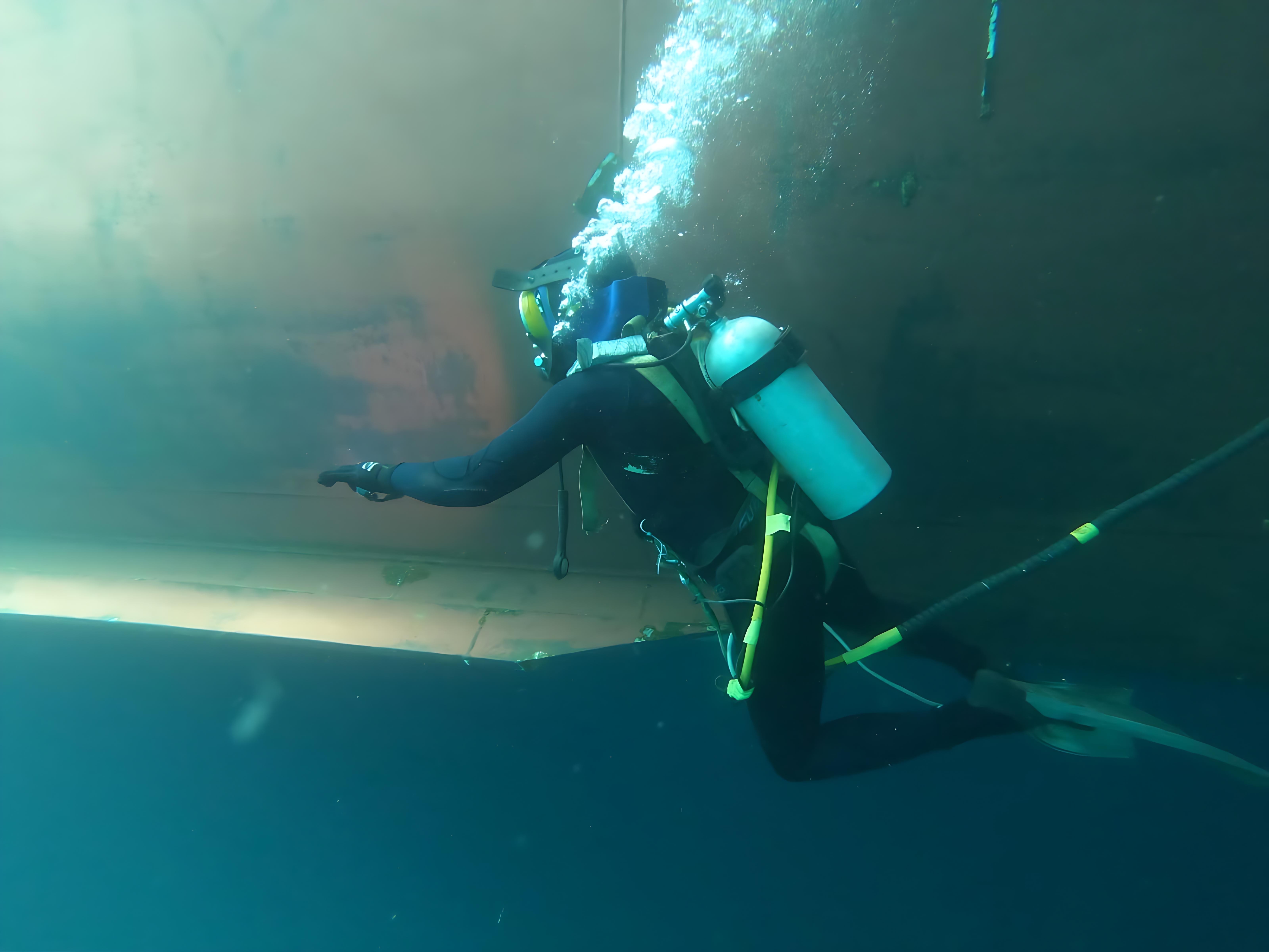 Seachios® Marine Services commercial diver conducting detailed underwater hull inspection along the flat bottom and bilge area of a trading vessel, visually checking weld seams, coating condition and marine growth as part of class-approved in-water survey and hull maintenance program.