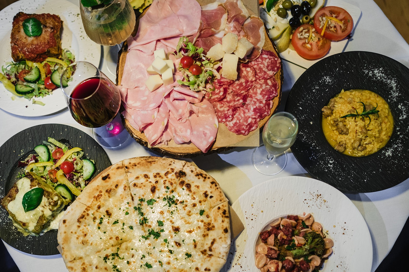 An overhead view of an Italian meal spread, featuring a black plate of saffron risotto with sausage, a charcuterie board with sliced cured meats and parmesan, a white plate of orecchiette pasta with vegetables and sausage, slices of pizza, fresh tomato salad, and a glass of white wine placed at the centre.