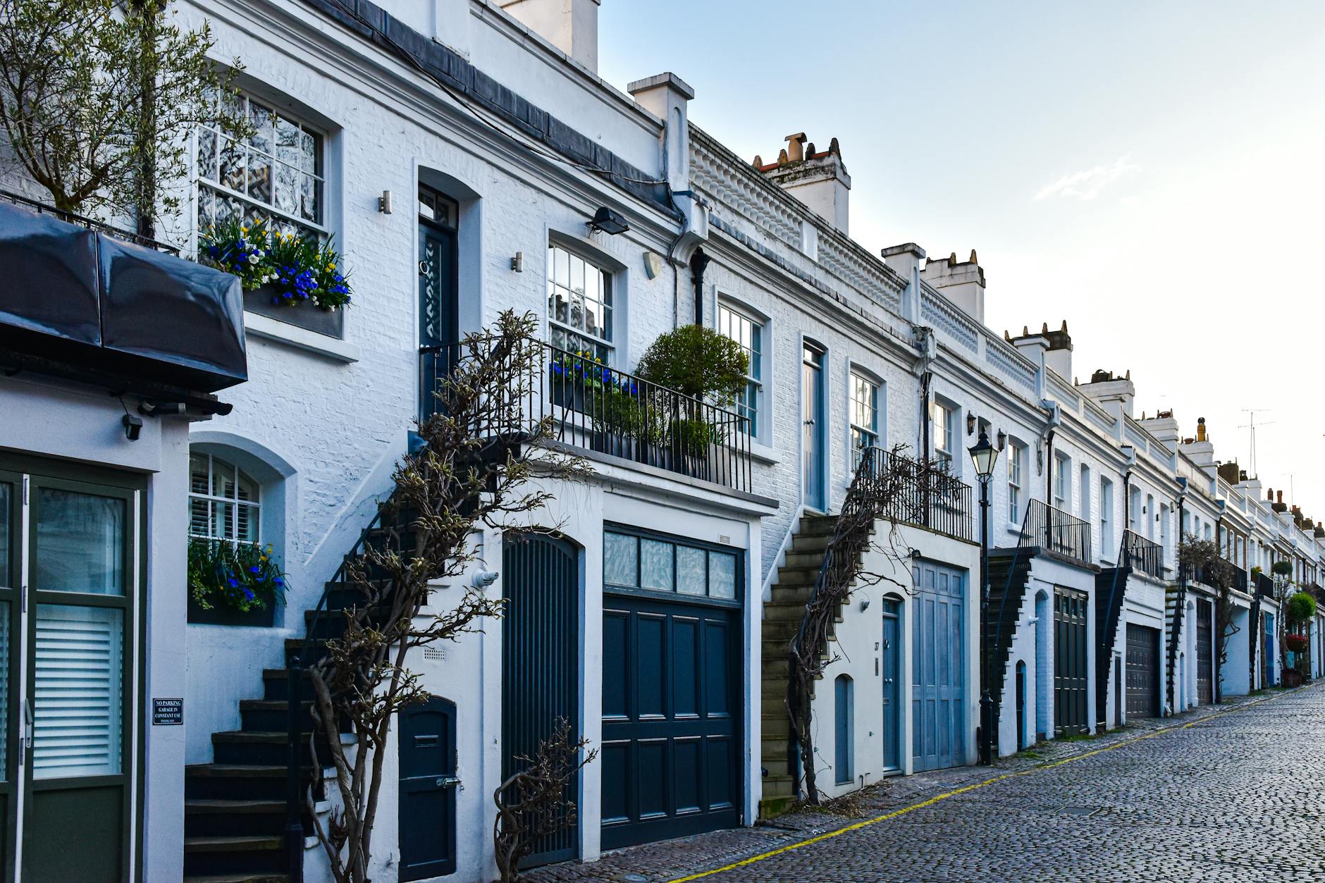 Row of white-painted Georgian mews houses with blue garage doors, iron staircases and window boxes on a cobblestone street