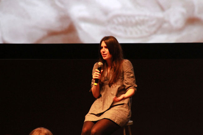 A woman with long brown hair holds a microphone in her right hand and gestures with her left hand while seated on a stool. She wears a light brown, loose-fitting dress and dark tights or stockings. A large projection screen behind her displays a close-up image that appears to show a face with teeth showing. The background is dark, and the image on the screen is out of focus. A portion of a person's head is visible in the lower left corner of the frame.