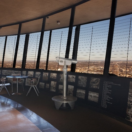 Observation deck with large windows, a telescope, and a small table with chairs. Cityscape visible through the windows.