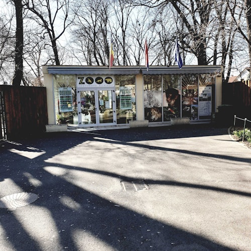 單層建築，配有大型窗戶，入口標示為「動物園」，頂部懸掛多面旗幟。背景中有光禿禿的樹木。