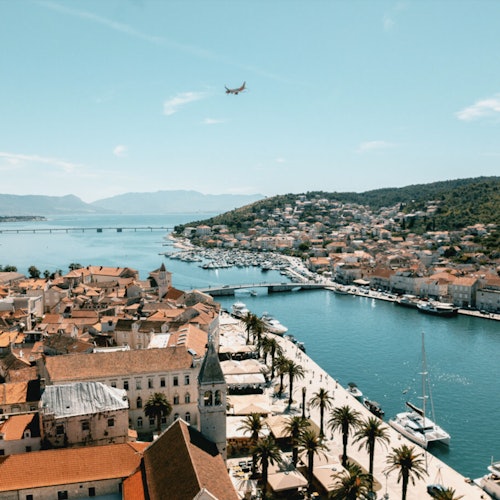 En kystby med rødtakede bygninger, en palmeprydet vannkant, fortøyde båter og et fly som svever over.