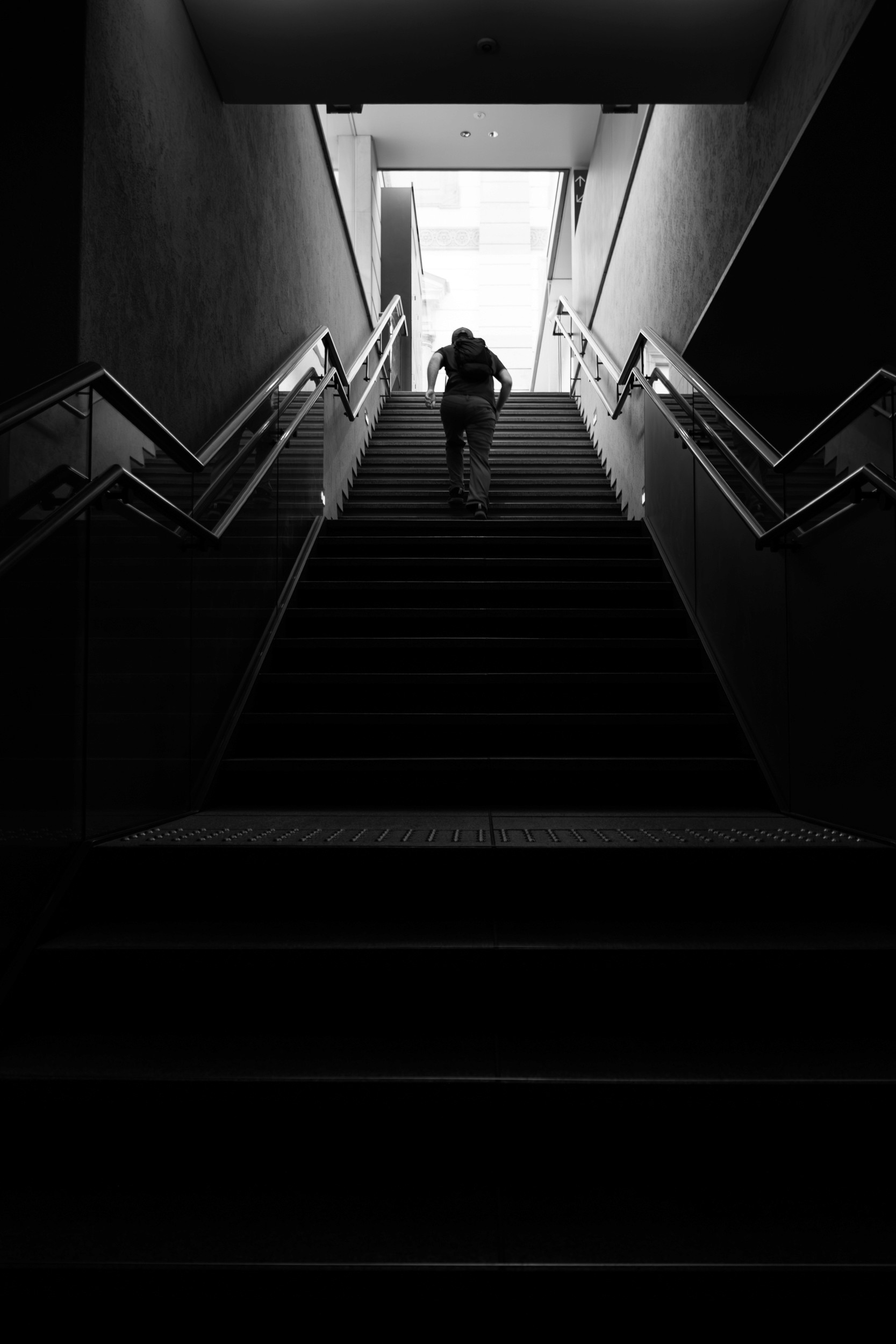 Black and white photograph looking down an escalator with a single person walking past bright light at the bottom of the frame.