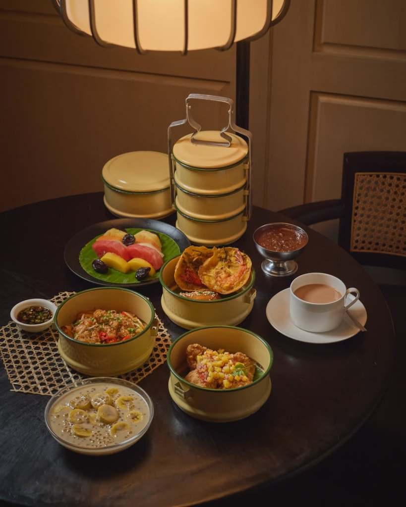 A table set with a variety of dishes including rice, fruits, and snacks, complemented by a cup of tea. The food is presented in stacked yellow tiffin containers and bowls, with a decorative placemat beneath.