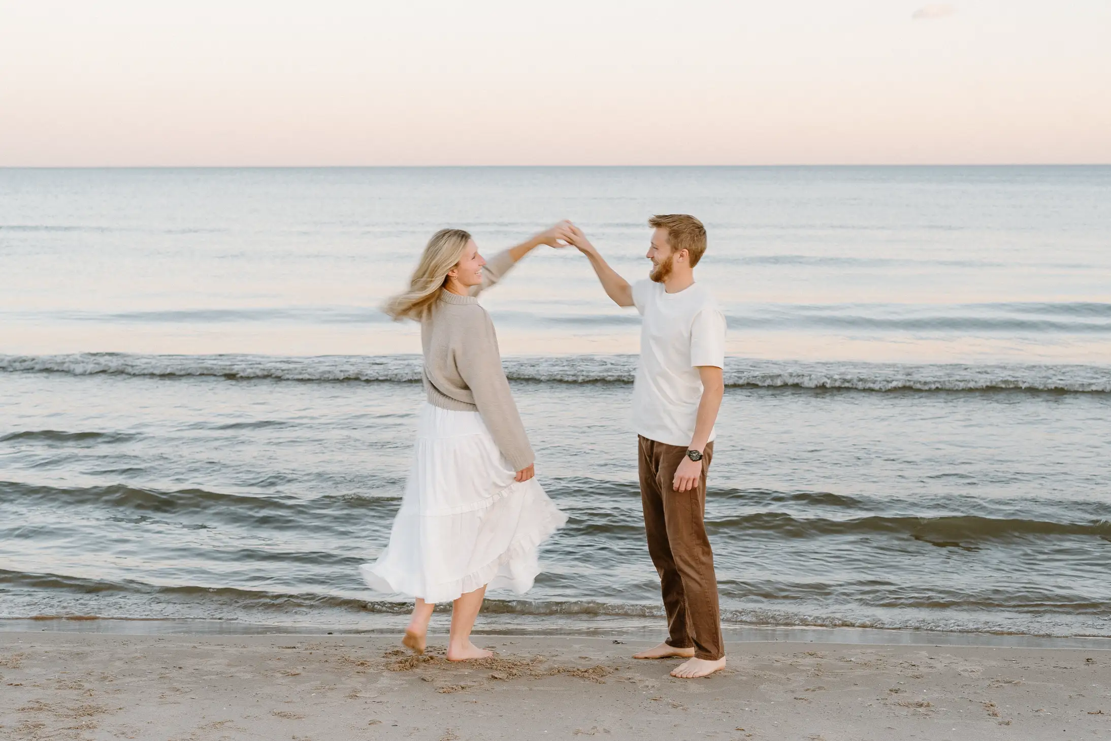 A couple dances hand-in-hand on the beach at sunset, with gentle waves in the background.