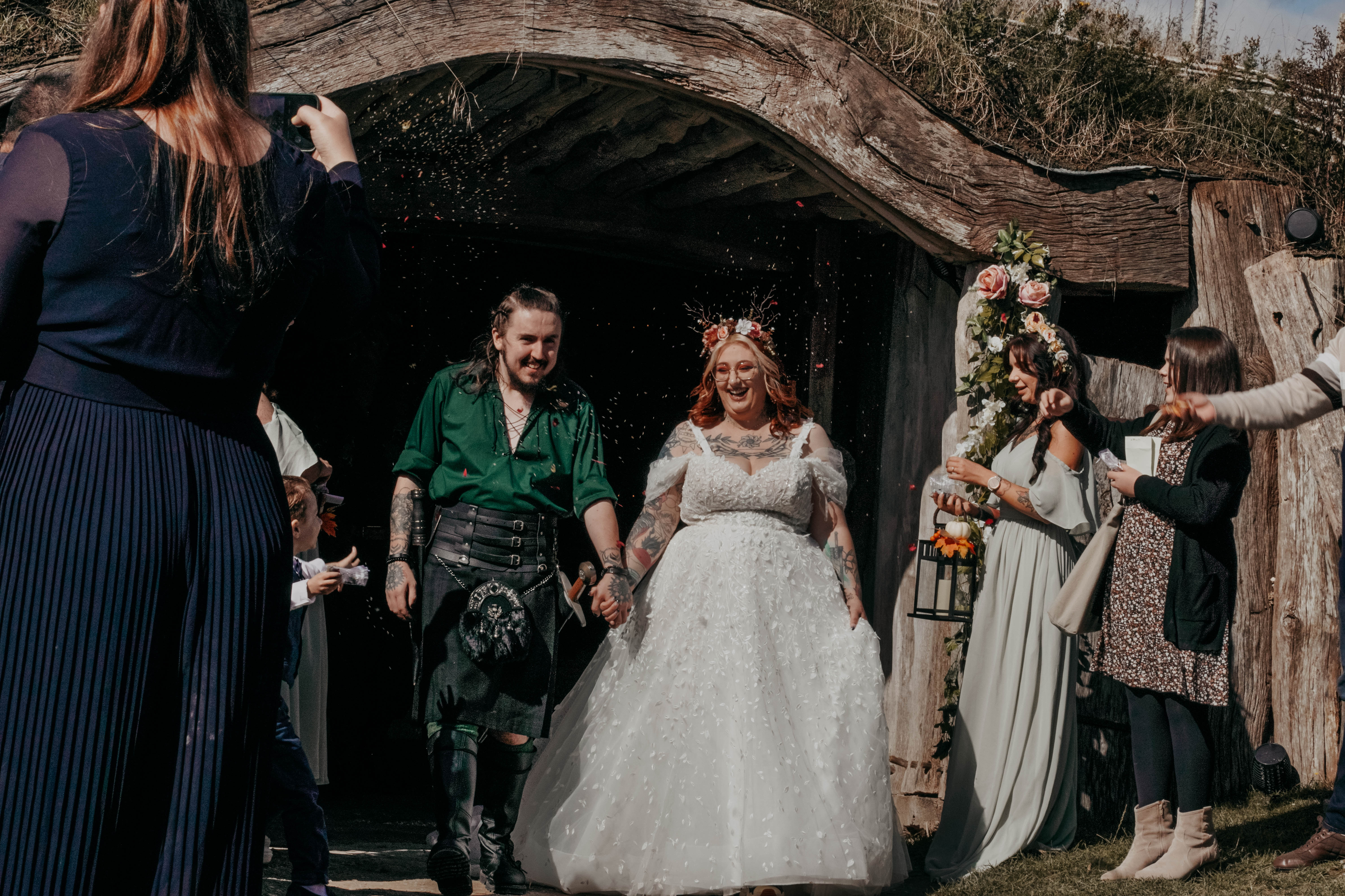 A smiling bride and groom walk hand-in-hand as guests throw confetti after their wedding ceremony. The groom has long hair and wears a green shirt and kilt, while the bride has red hair, a floral crown, and a white gown. They are exiting a rustic building with a curved, natural wood entrance.