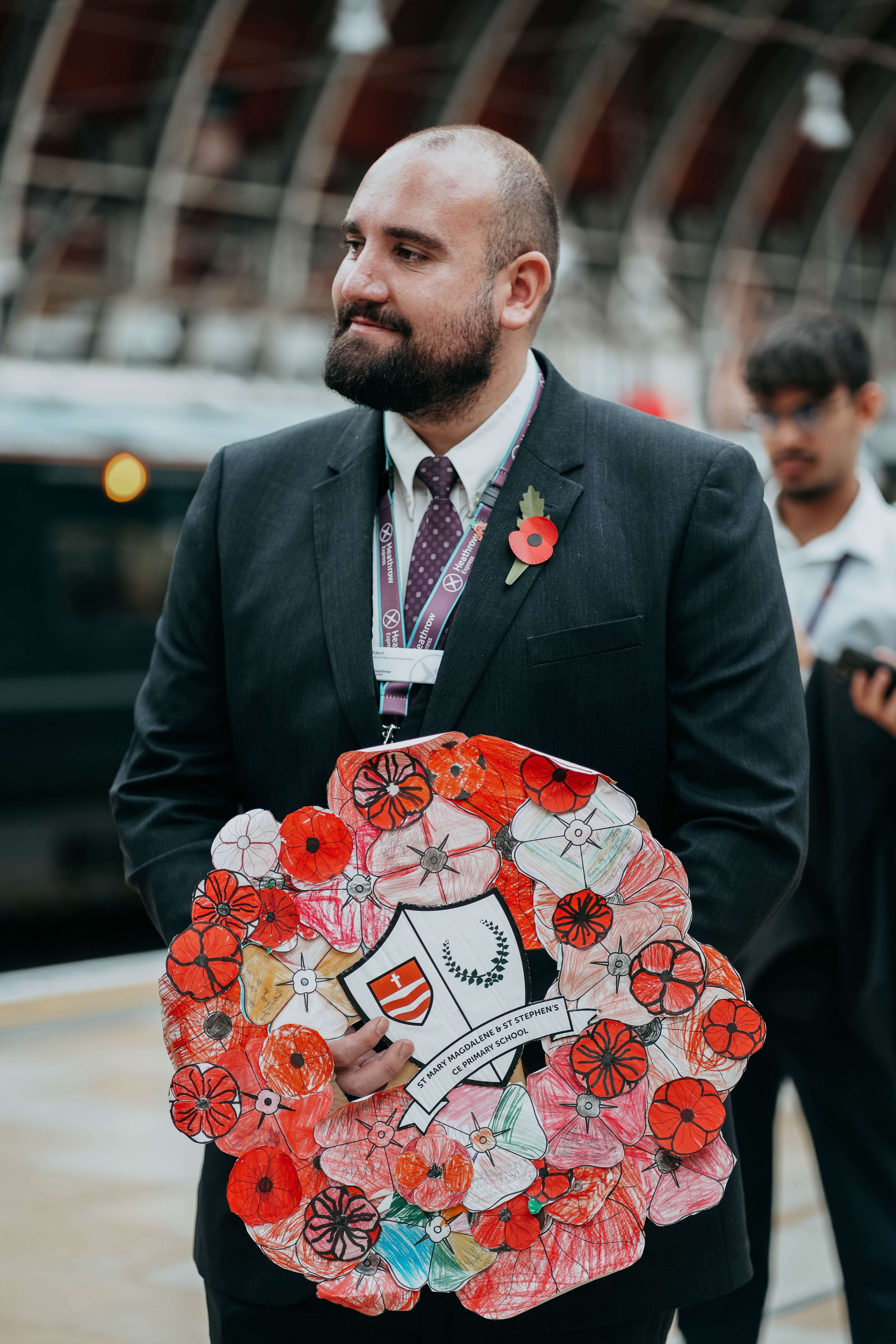 A man in a suit with a poppy on his lapel stands on a train platform, proudly holding a large, handmade wreath made of colorful, child-drawn paper poppies.
