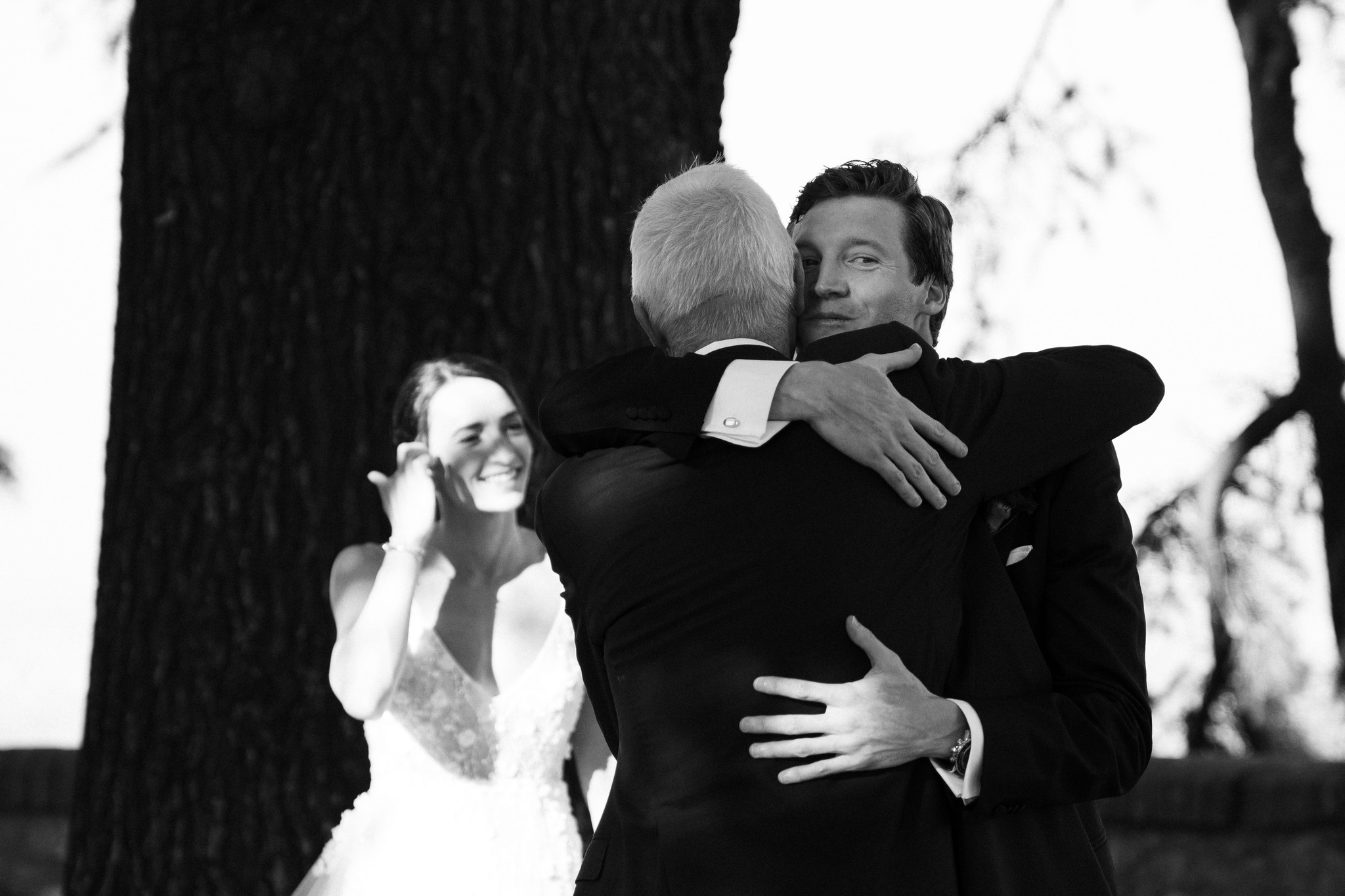 Lo sposo abbraccia un ospite sotto gli alberi di Tabiano, la sposa osserva: momento emotivo del reportage matrimoniale