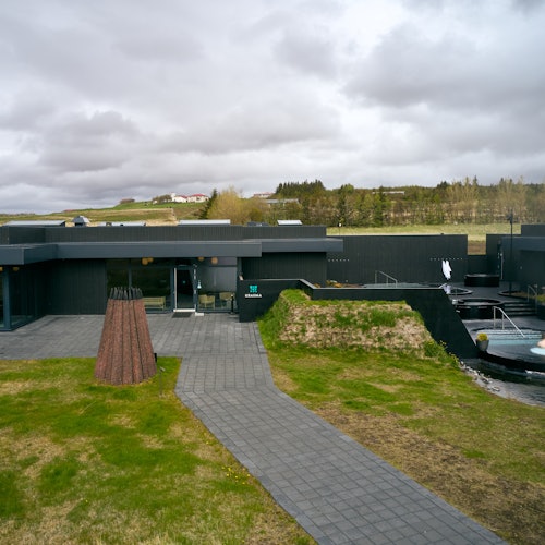 Dark modern building with large windows, surrounded by grassy area and walkway, under a cloudy sky, with hilly terrain in the background.