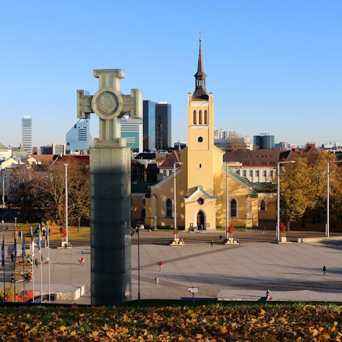 A győzelem oszlopa mögött, a város főterén, magas toronnyal rendelkező sárga templom áll, a háttérben modern épületekkel.