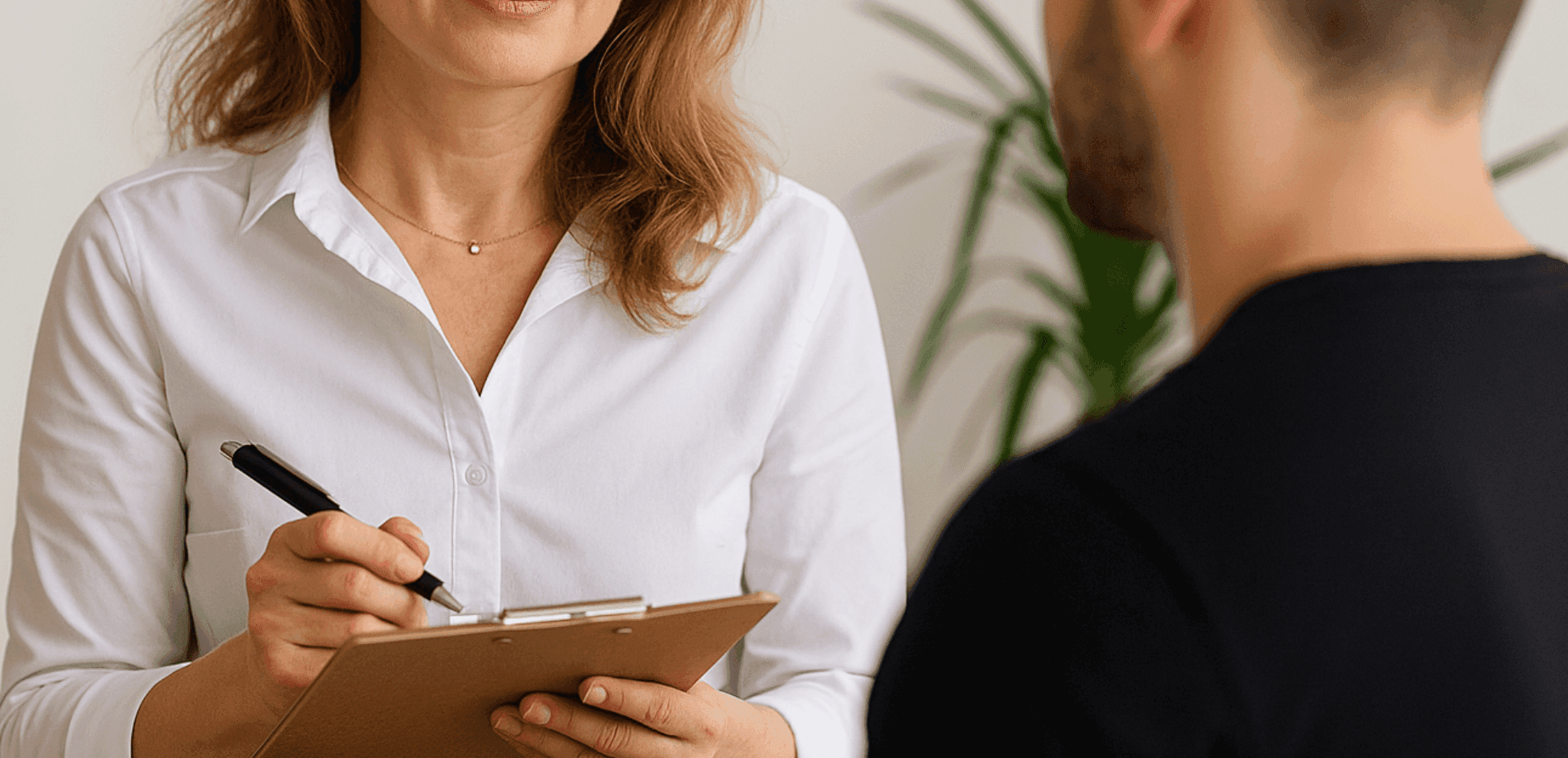 A woman in a white shirt holds a clipboard, talking to a man in a black shirt, with plants in the background.