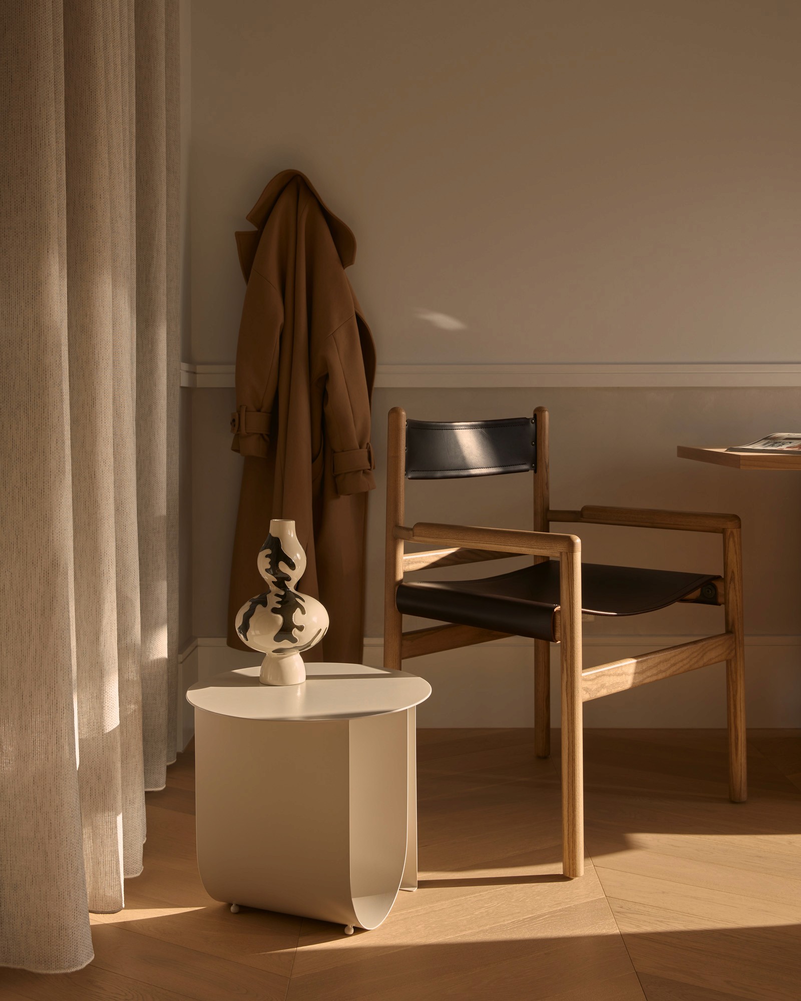 Wooden armchair with black leather seat and back beside a side table with a ceramic vase, a camel coat hanging on the wall behind