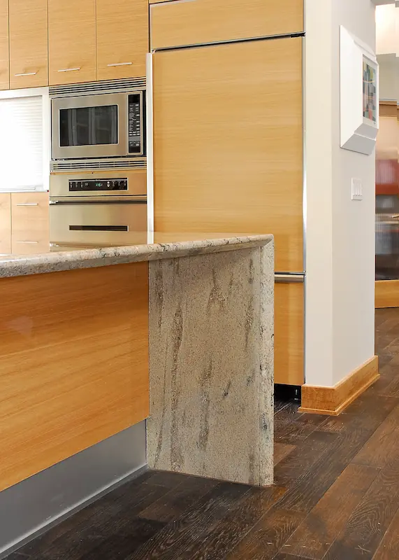 Close up of quartzite island countertop waterfall edge.Modern kitchen with wooden cabinets, central island, hanging lights, and dark flooring. Irvine Transitional Modern. Photo by Chris Darnall.