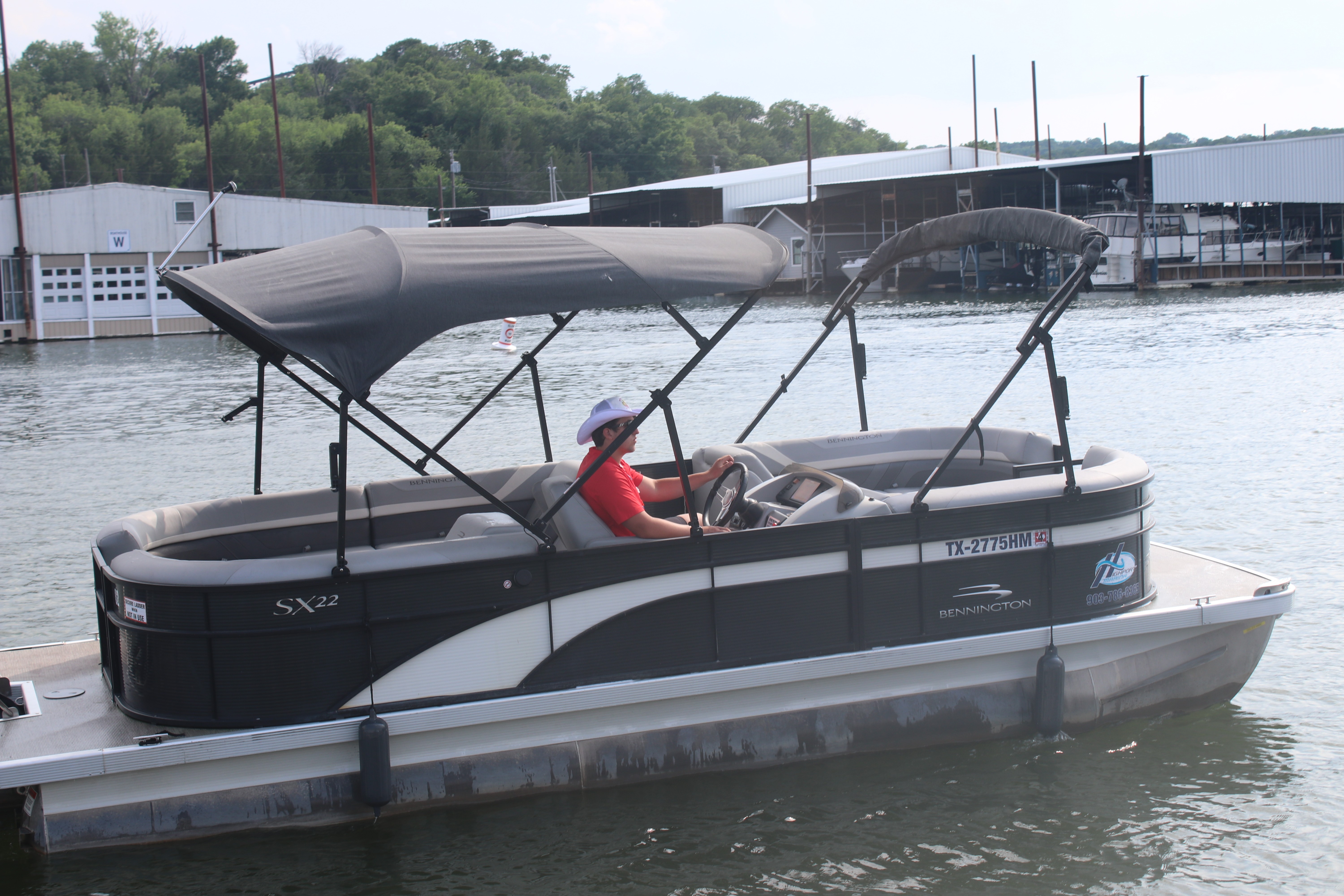 A person is driving a pontoon boat with a black canopy on a calm lake, with boathouses and lush green trees in the background.