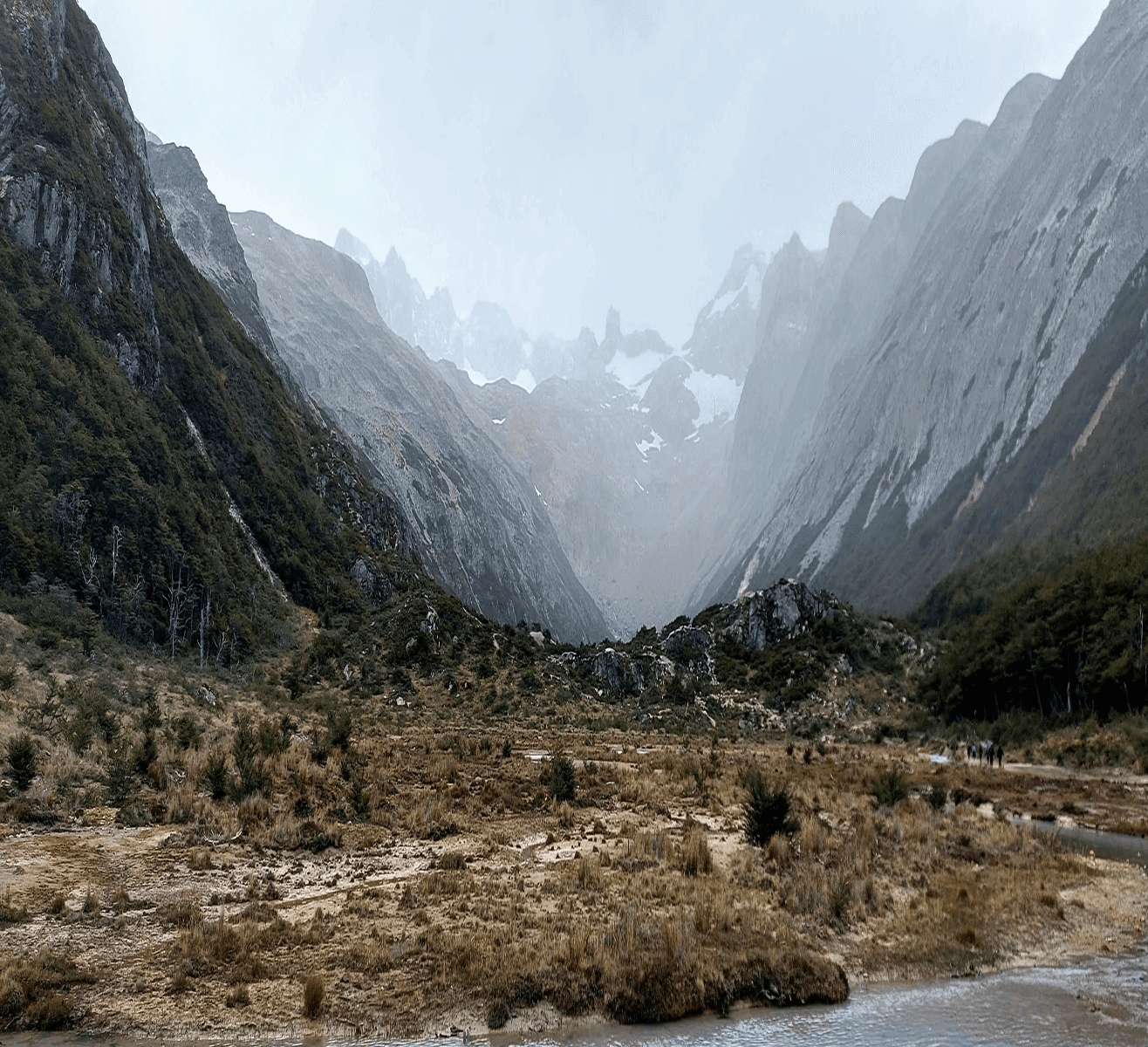 Rugged valley with sparse vegetation leading to jagged, mist-shrouded mountain peaks.