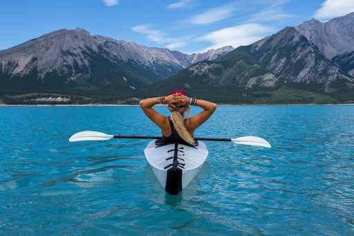 A person relaxes in a kayak on clear blue water with mountains in the background under a bright sky.
