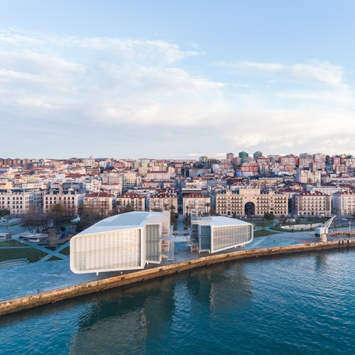 Modern waterfront buildings near a cityscape with densely packed buildings, a park area, and a clear blue sky.
