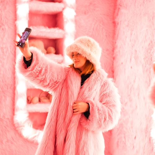 Persona con cappotto e cappello di pelliccia rosa che si scatta un selfie in una stanza con pareti e decorazioni soffici e rosa.