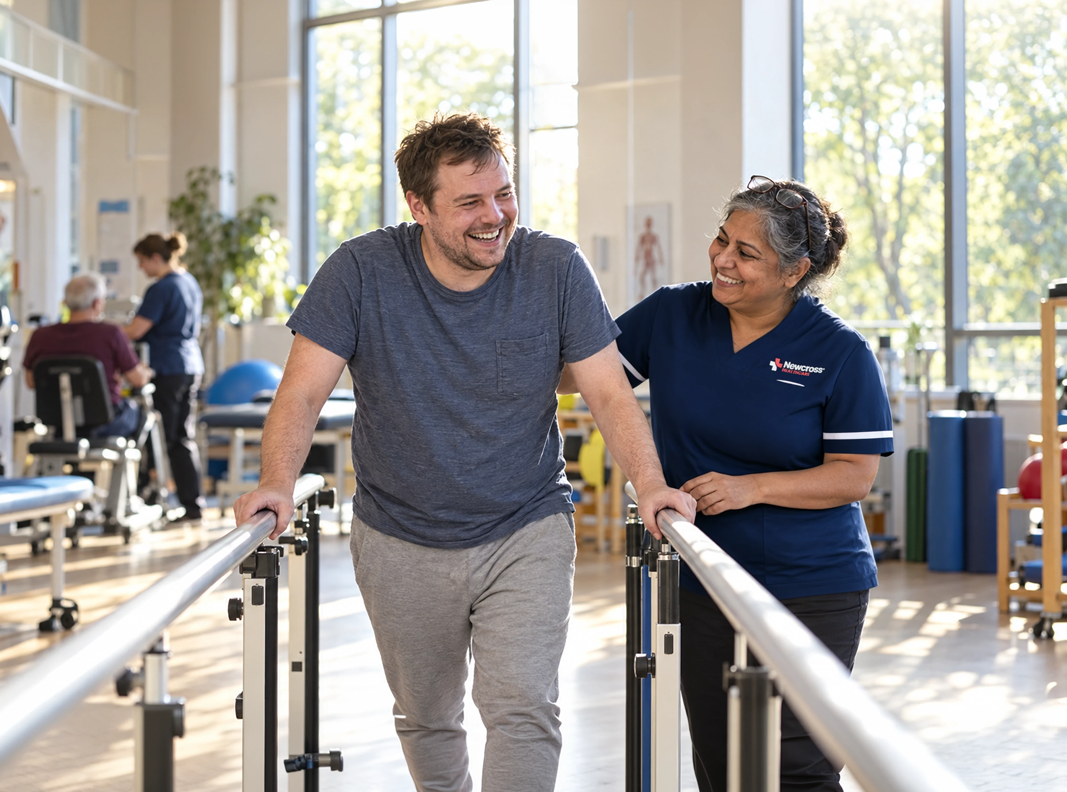 Man recovering from a brain injury smiles while practising walking between parallel bars with support from a physiotherapist in a bright neurological rehabilitation unit.