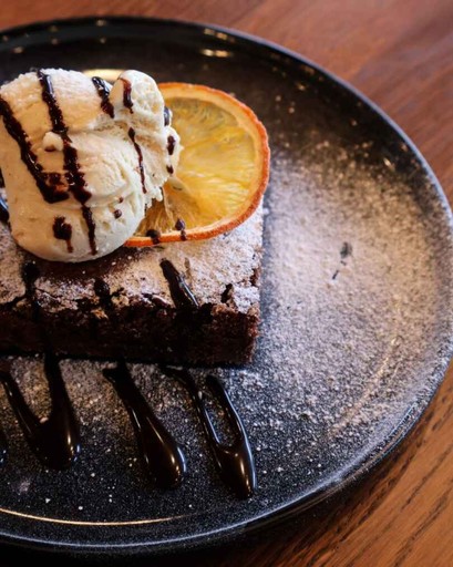 Rich chocolate brownie topped with vanilla ice cream, an orange slice, and chocolate drizzle on a black plate.