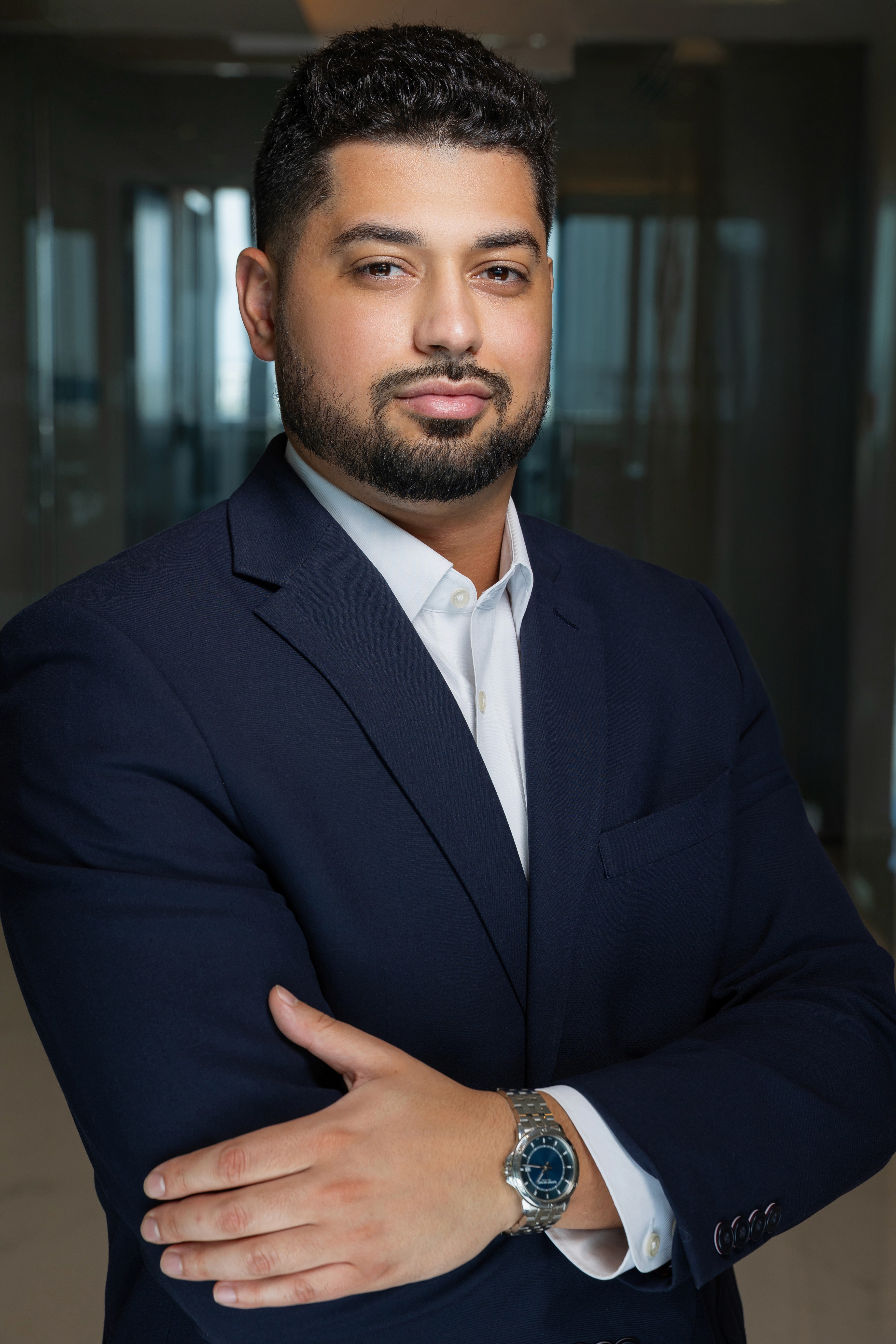 A woman in a black blouse and grey trousers stands confidently with arms crossed, in front of wooden shelves with decorative pottery in a modern office.