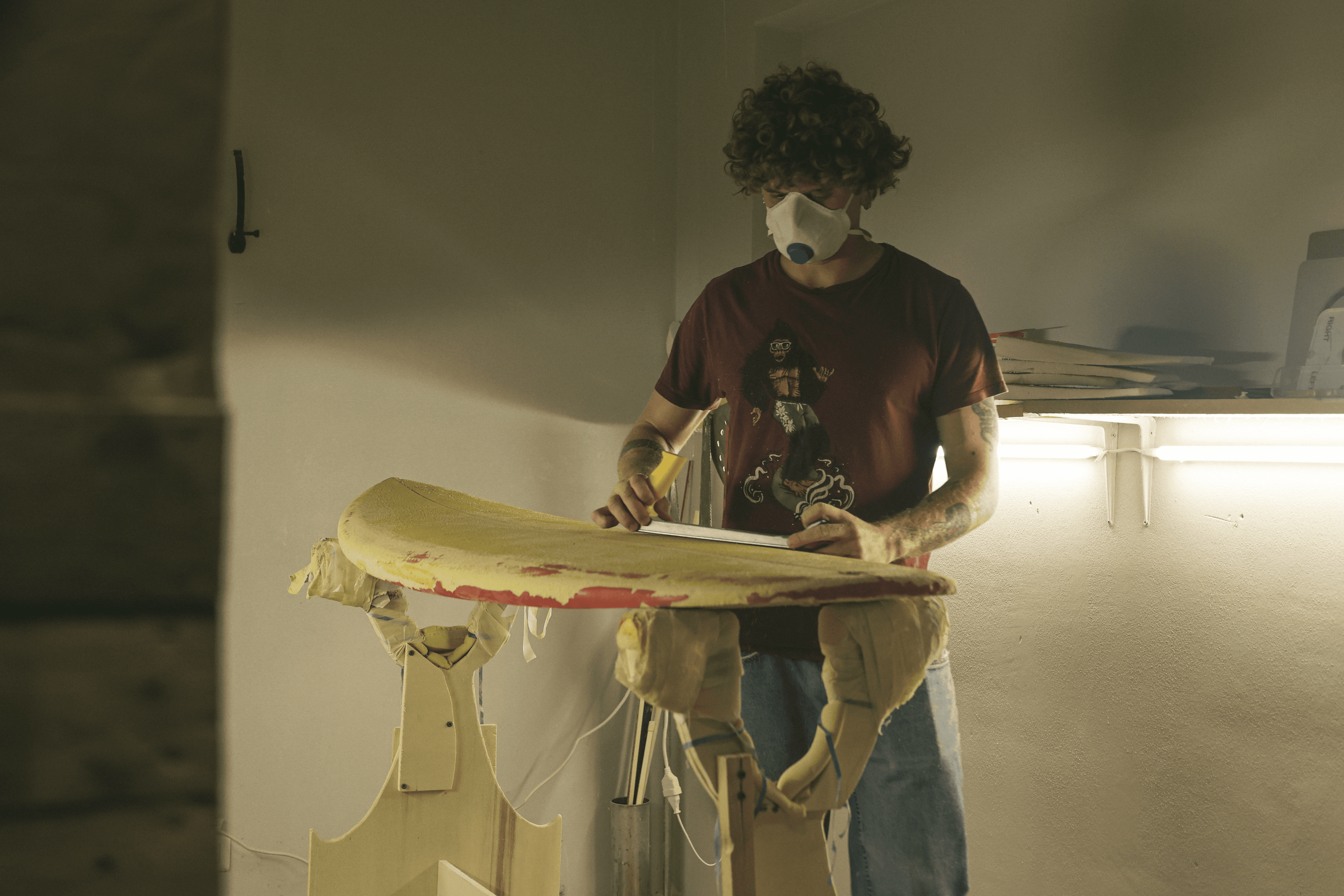 Shaper con mascherina protettiva e maglietta bordeaux, lavora sul contorno di una tavola da surf gialla e rossa su un cavalletto in laboratorio.