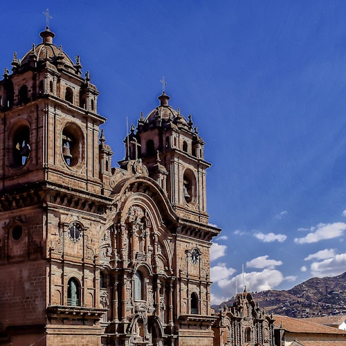 一座歷史悠久的石教堂，兩座鐘樓矗立在澄藍的天空之下，背景是山脈。