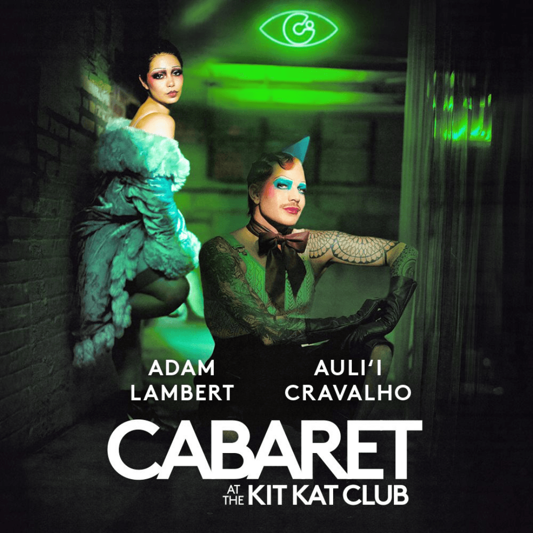 Cabaret at the Kit Kat Club on Broadway. In a green lit alley with the Kit Kat Club logo in a neon light in the top right.  Gayle Rankin (as Sally Bowles) leans against the wall, one foot against the wall. Eddie Redmayne (as the Emcee) squats in the center of the alley in a puddle. He is topless, wearing a tie and a birthday hat.