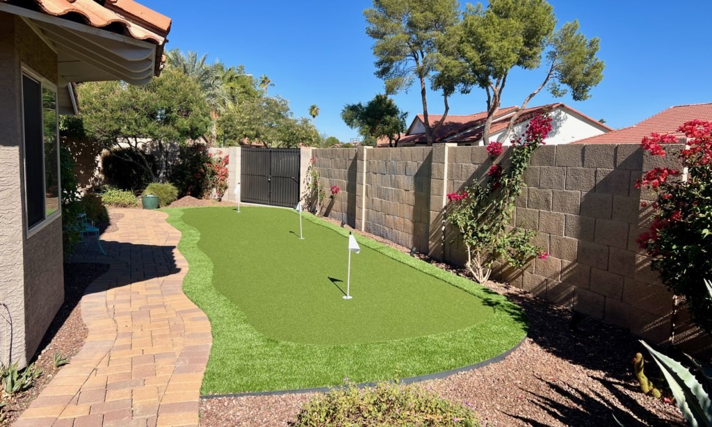 The image shows a beautifully landscaped backyard featuring a brick pathway and a well-maintained, small artificial putting green, surrounded by an adobe-style fence, lush greenery, and vibrant flowering plants, offering a glimpse of an affordable luxury vacation rental house experience.