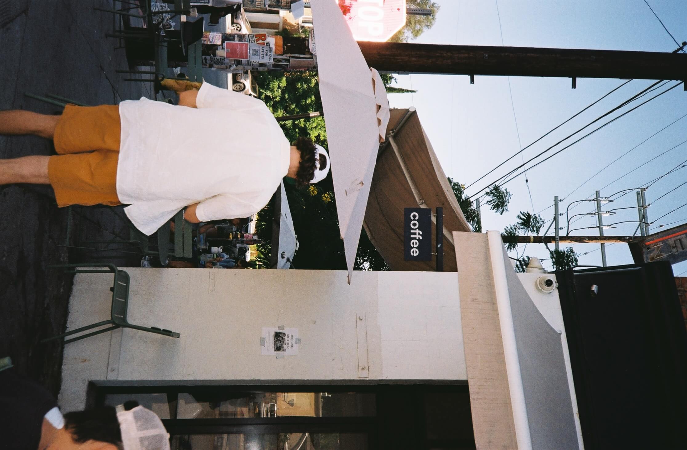 A man in a white shirt and terracotta shorts viewed from behind walking towards a cafe