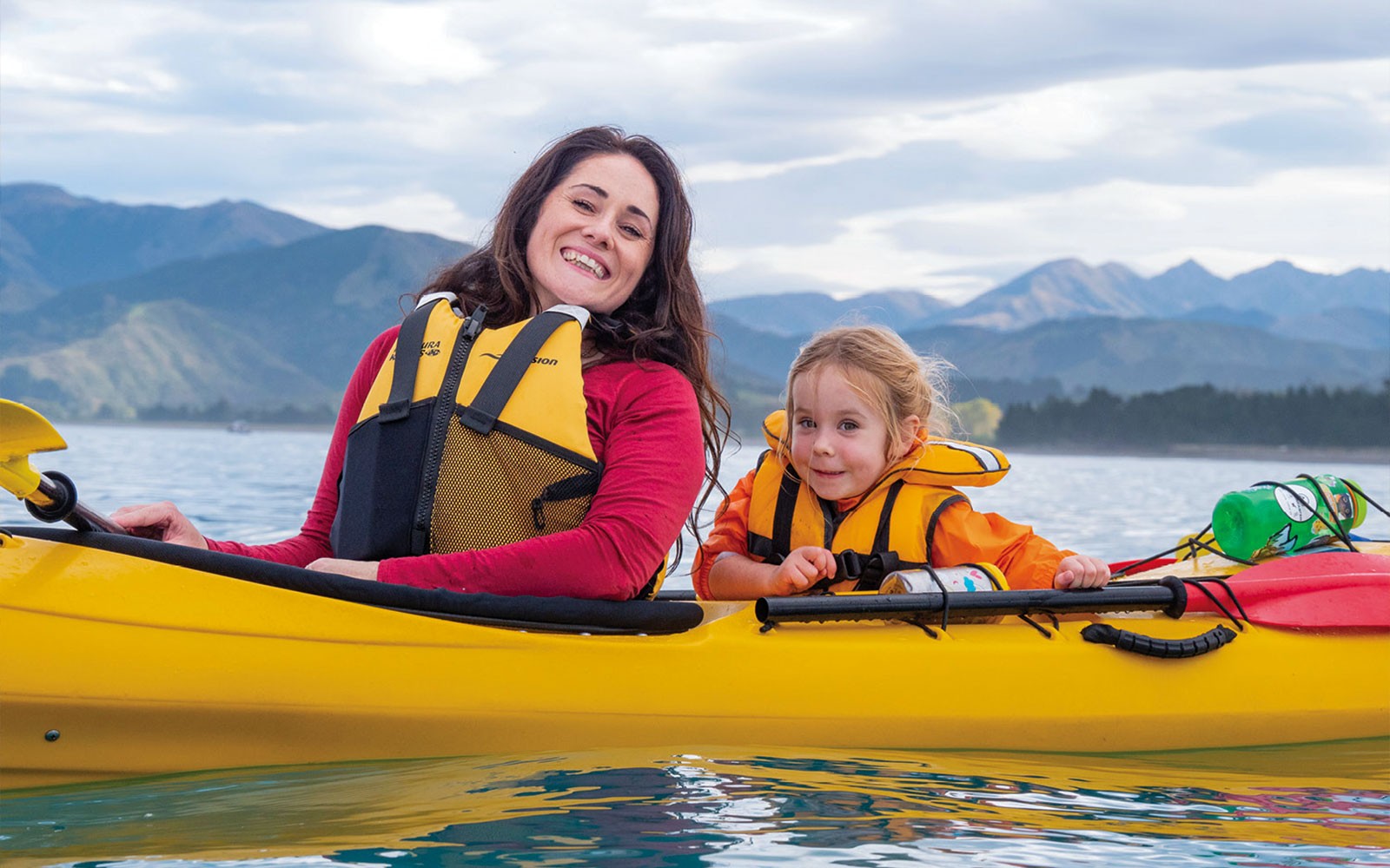 Mutter und Kind beim Kajakfahren auf einem See mit Bergen im Hintergrund.