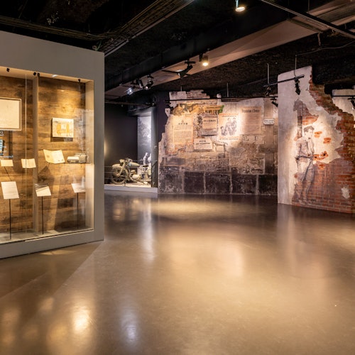 En museiutställning med en monter som innehåller dokument och artefakter, samt ett omgivande område med väggmålningar och en veteranmotorcykel.