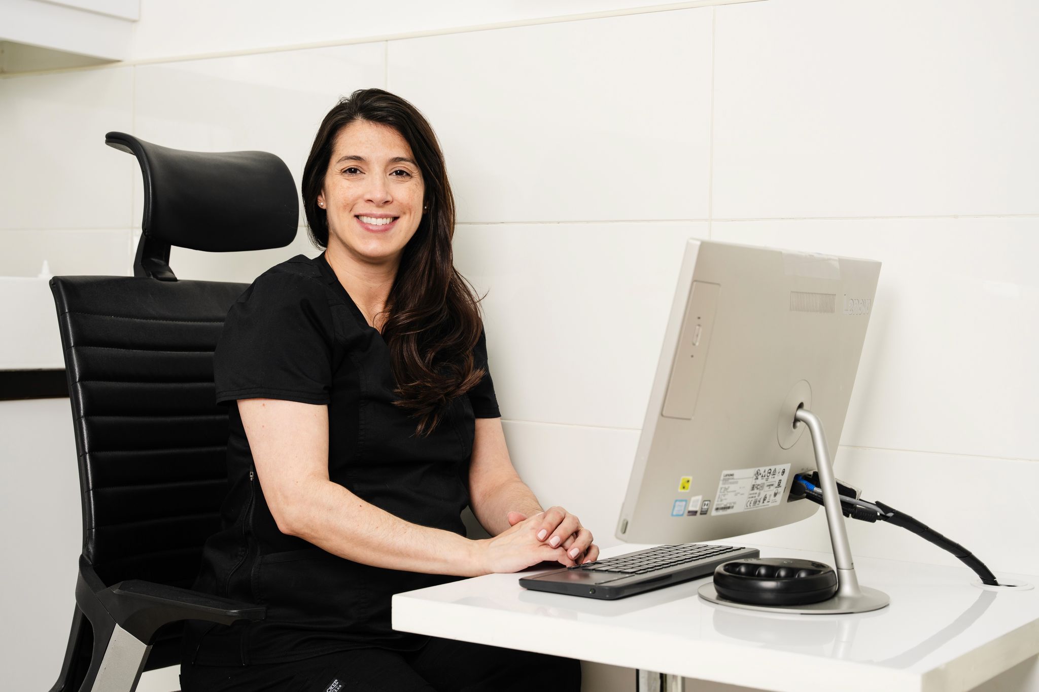 A smiling woman in black medical scrubs sits at a desk in a modern, white clinical office. She is seated in a black ergonomic chair and working on a desktop computer. The setting appears to be at Noble Vet Clinics. 