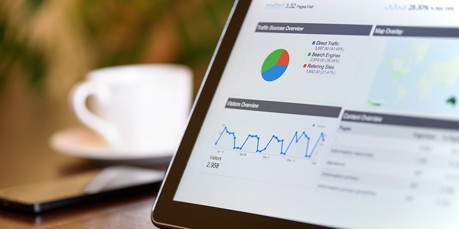 Close-up of a tablet screen showing digital marketing analytics, including traffic sources and visitor overviews. The tablet sits on a dark wood table in a cafe-like setting with a blurred coffee cup in the background.