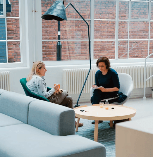 A woman lying down on a soft surface with her eyes closed, bathed in warm sunlight filtering through leaves. Her face has a natural glow, reflecting relaxation and tranquility in a serene spa-like setting.